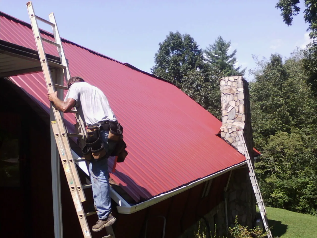 Roofer on a ladder beside a red metal roof and stone chimney — Soffit & Fascia Repair by Roofing Brothers in Hinckley